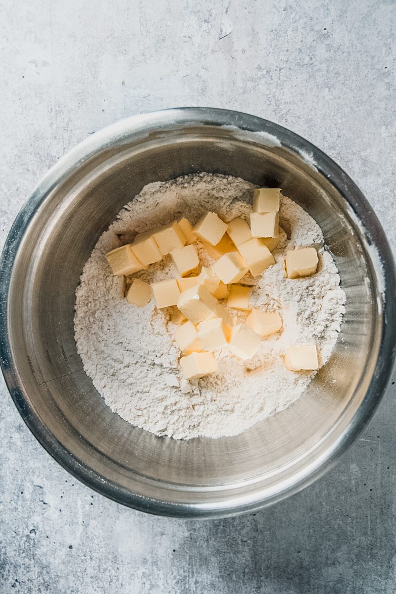 Cubes of butter sit on top of flour in a stainless steel bowl on a gray surface.