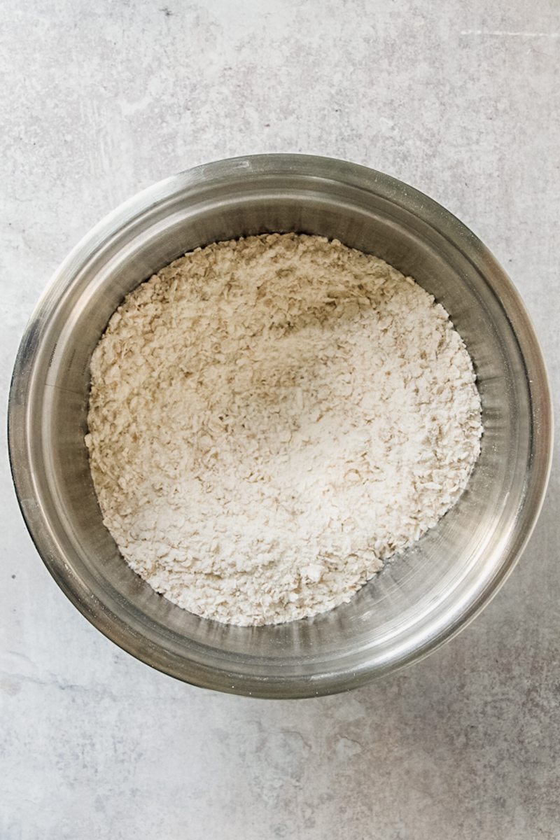 A dry Anzac biscuit mixture sits stirred in a stainless steel bowl on a gray surface.