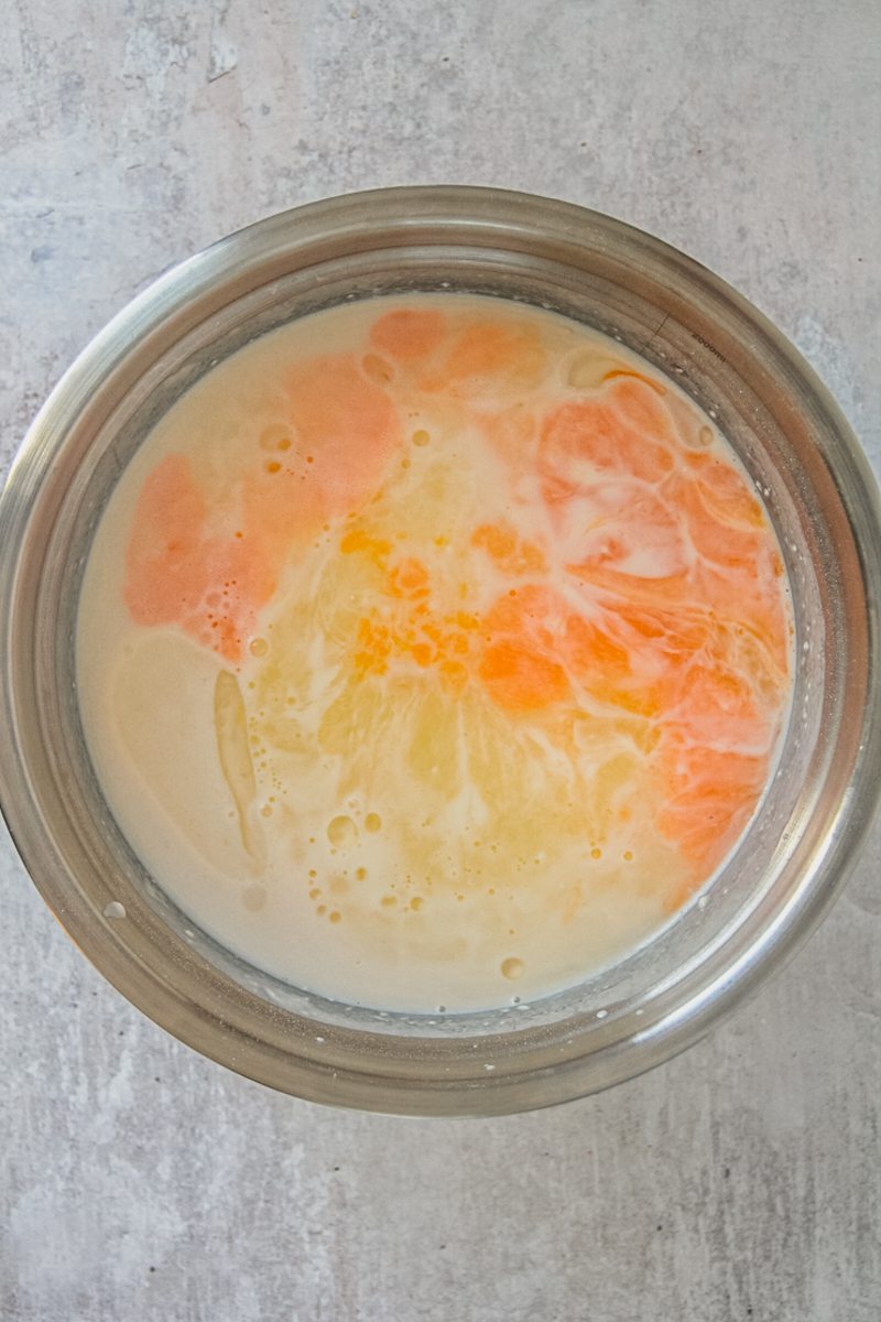 Eggs and oil sit on top of a loose pancake batter in a stainless steel bowl on a gray surface.