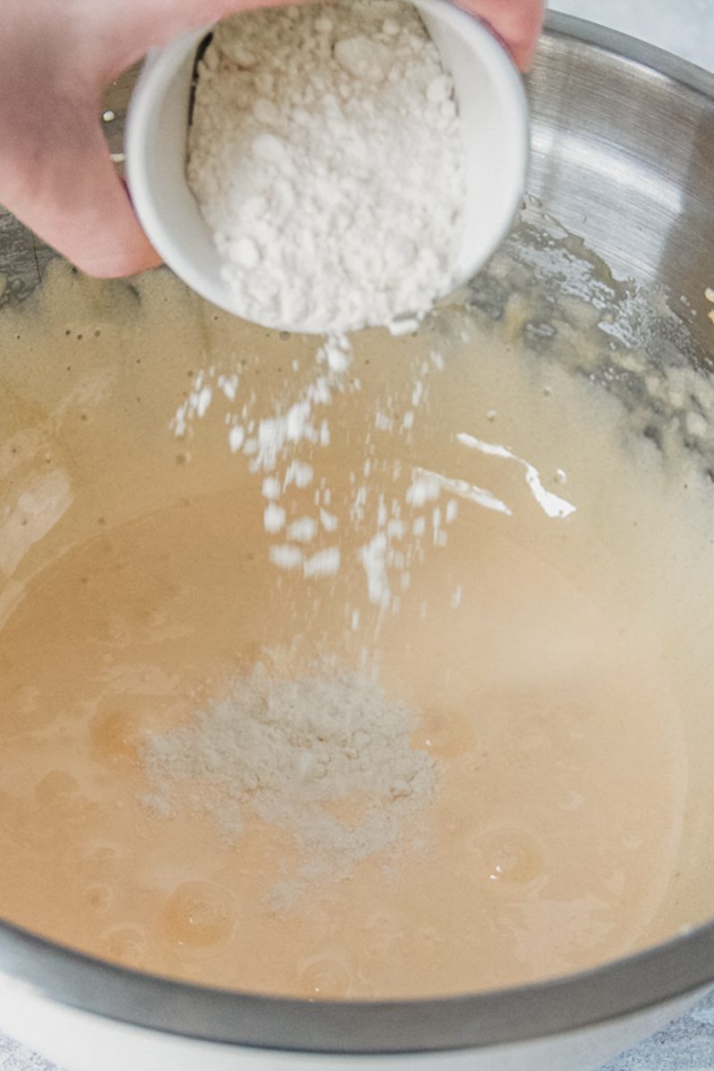 Flour is tossed over whipped egg yolks in a stainless steel bowl on a gray surface.