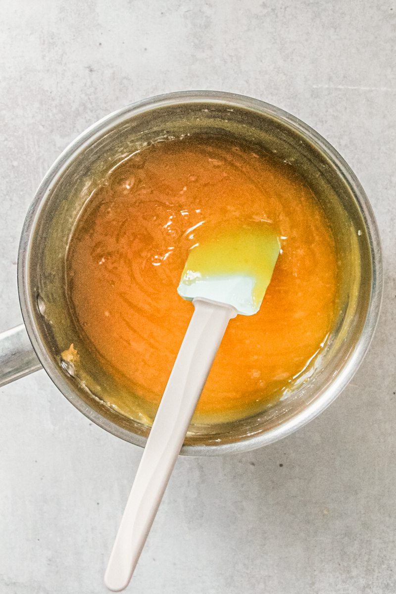 A lemon curd mixture sits in a stainless steel saucepan with a spatula resting in the center on a gray surface.