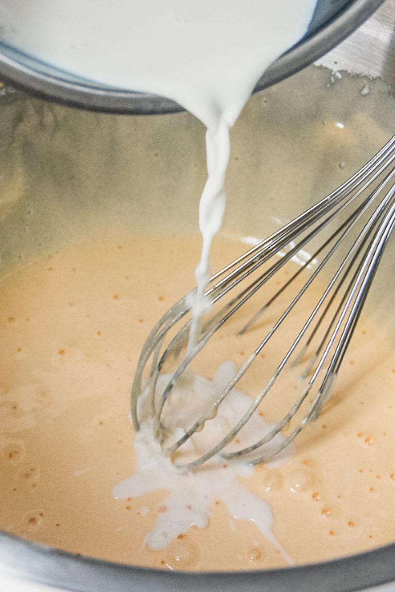 Milk is poured into a base custard recipe in a stainless steel bowl with a whisk.