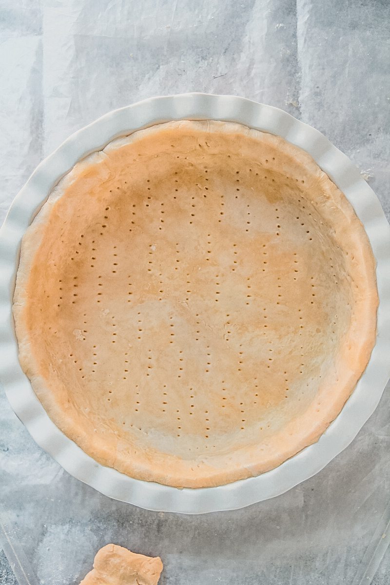 Unbaked pie pastry sits pierced in a white ceramic pie dish on a gray surface.