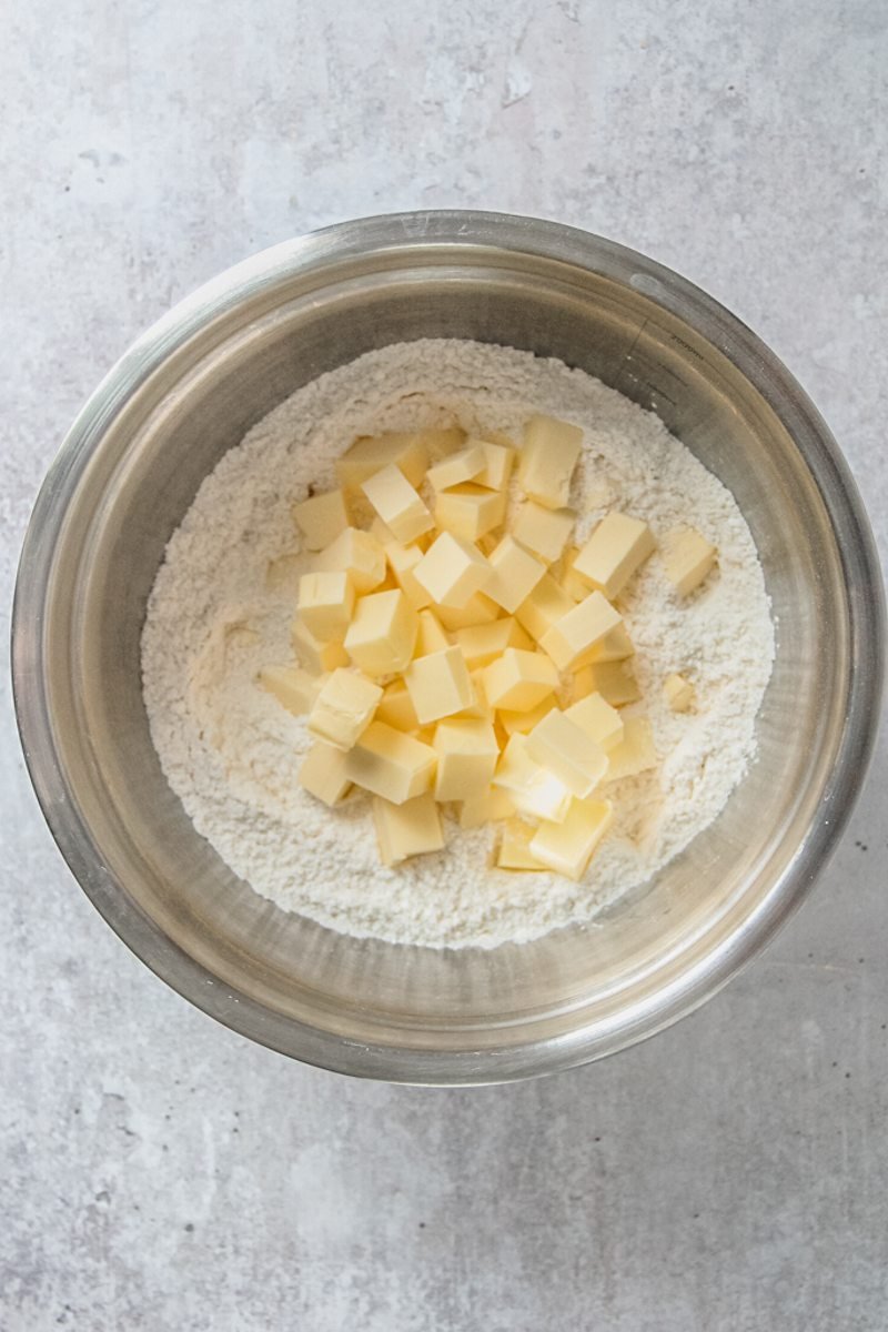 Cubes of butter sit on top of a flour mixture in a stainless steel bowl on a gray surface.