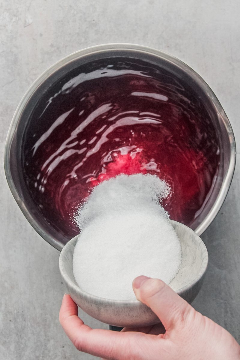 Sugar is poured into a cranberry juice mixture in a stainless steel saucepan on a gray surface.