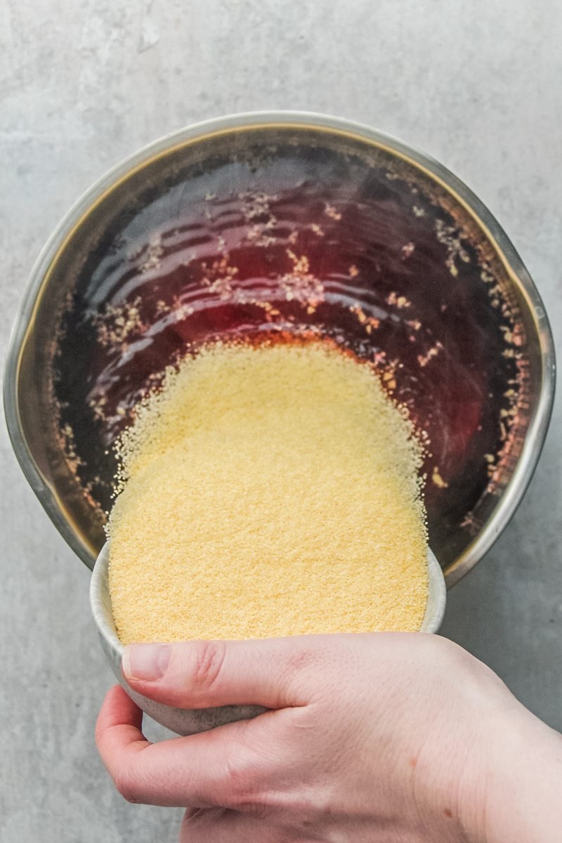 Semolina is poured into a cranberry mixture in a stainless steel saucepan on a gray surface.