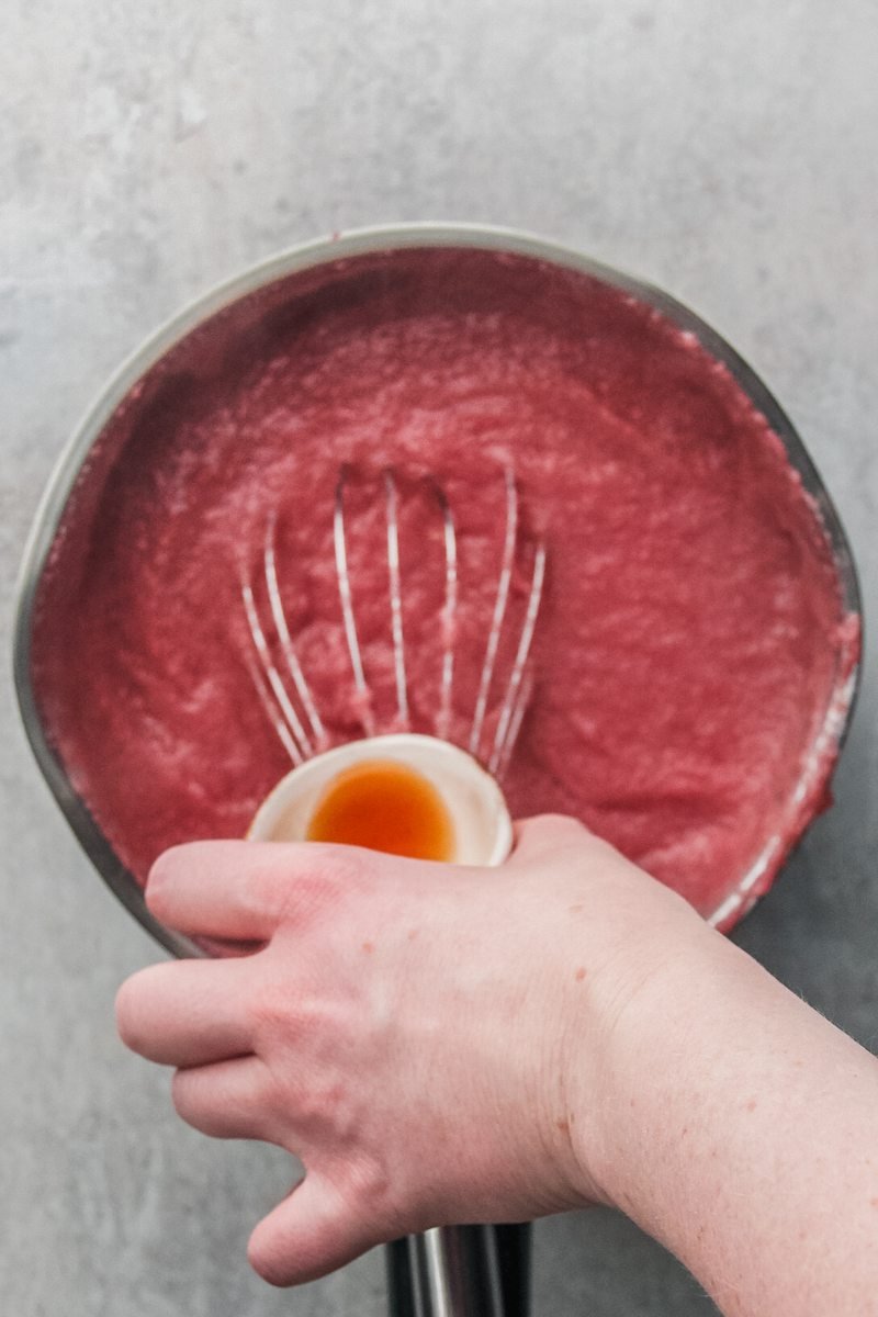 Vanilla extract is poured into a cranberry semolina pudding in a stainless steel saucepan on a gray surface.