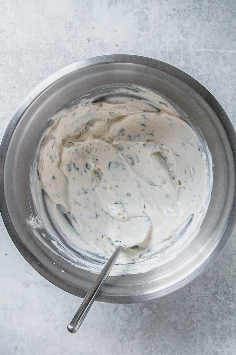 A herbed cream cheese mixture sits in a stainless steel bowl on a gray surface.