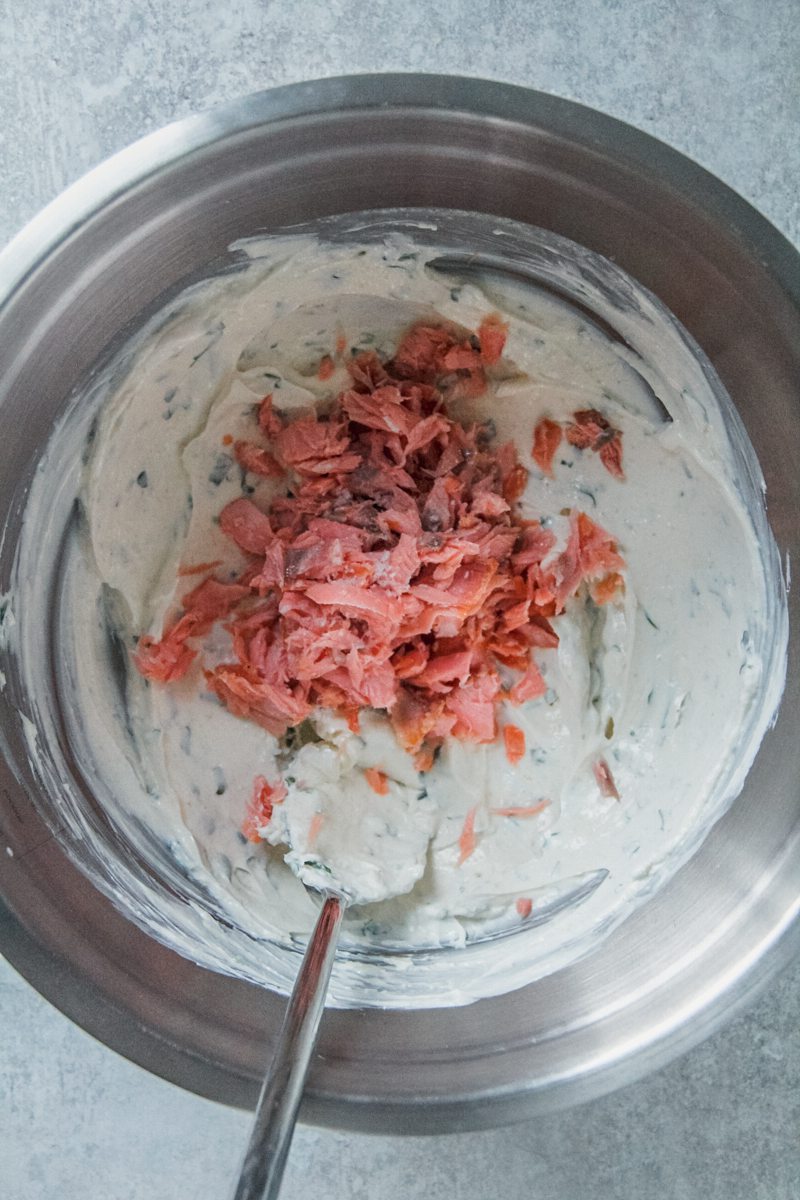 Flaked smoked salmon sits on top of a herb cream cheese mixture sits in a stainless steel bowl on a gray surface.