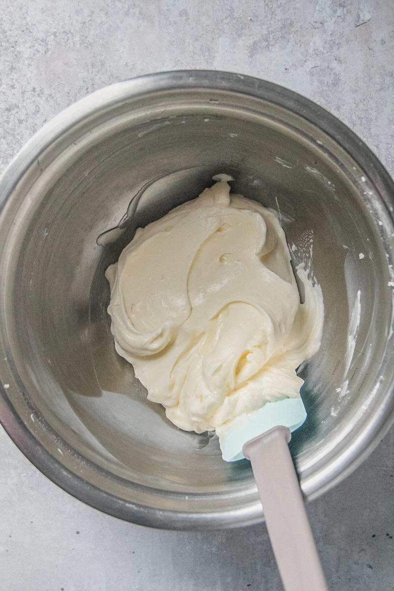 Smooth cream cheese frosting sits in a stainless steel bowl on a gray surface.