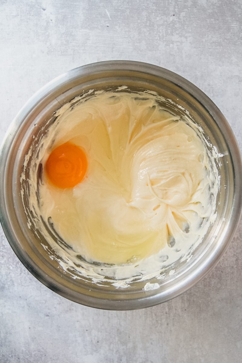 Whipped butter, sugar and an egg sit in a stainless steel bowl on a gray surface.