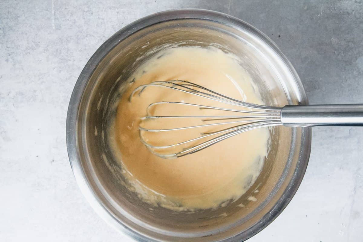 Pale custard sits in a stainless steel bowl with a whisk on a light grey background.
