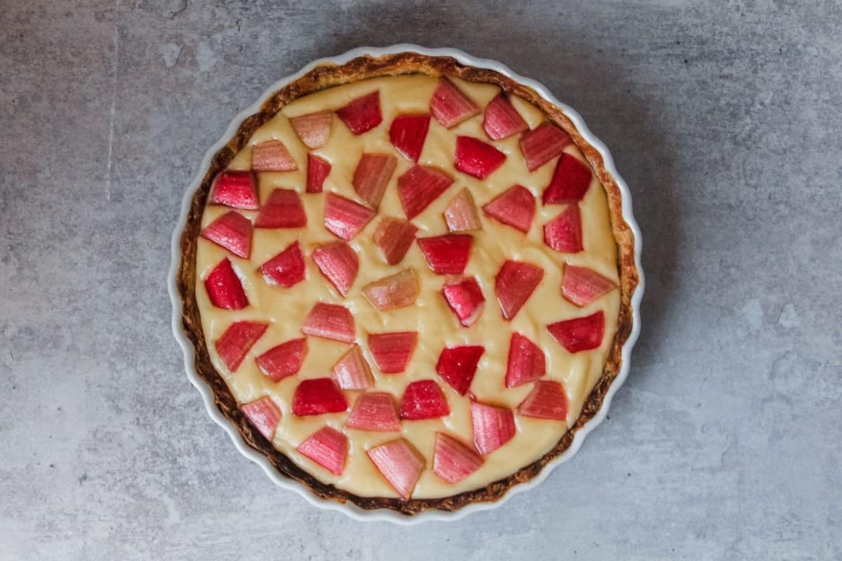 A rhubarb custard tart sits on a grey background prior to baking with bright pieces of rhubarb placed into a lemon custard.
