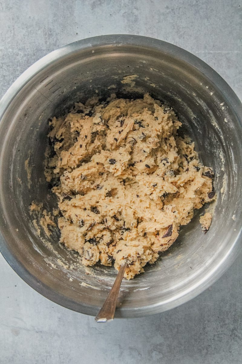 Chocolate chunk cookie dough sits in a stainless steel bowl on a gray surface.