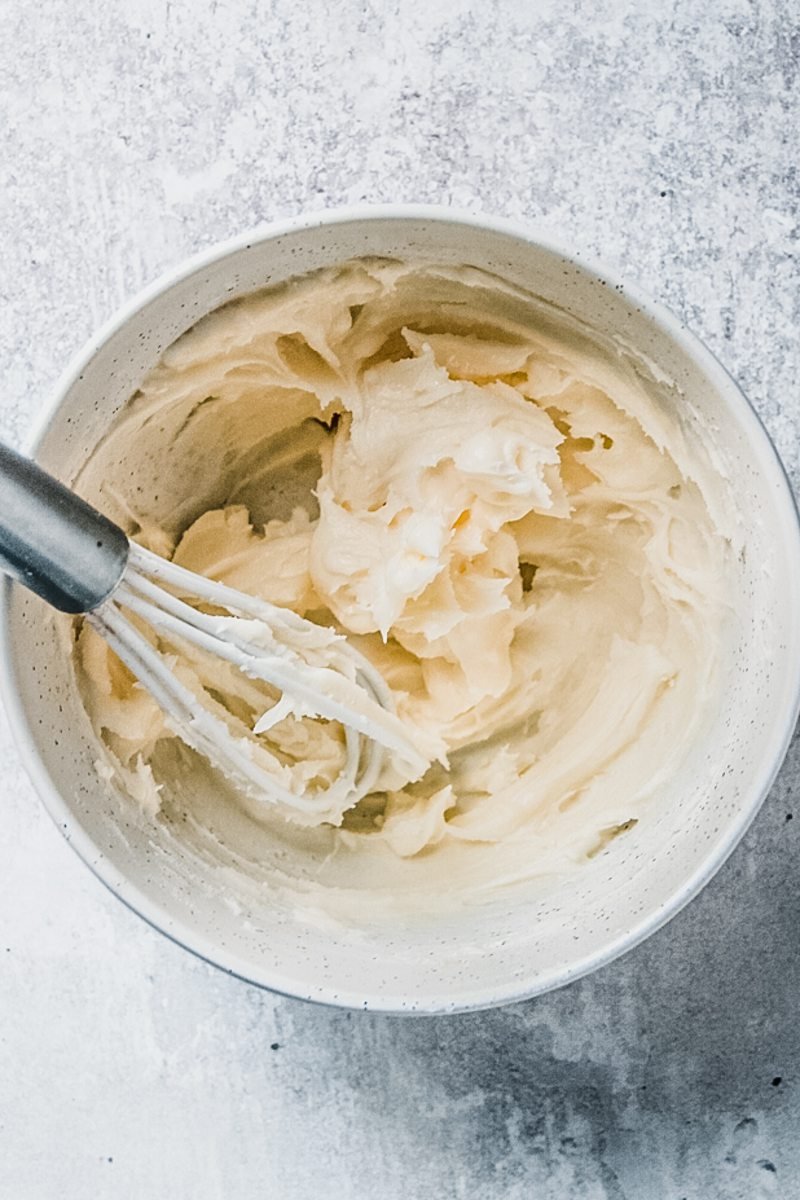 A milk frosting sits in a white ceramic bowl with a whisk on a gray surface.