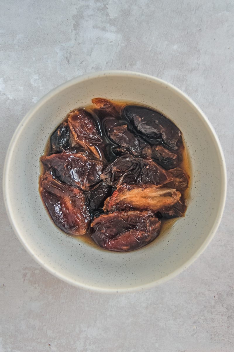 Seedless dates sit covered in boiling water in a white ceramic bowl on a gray surface.