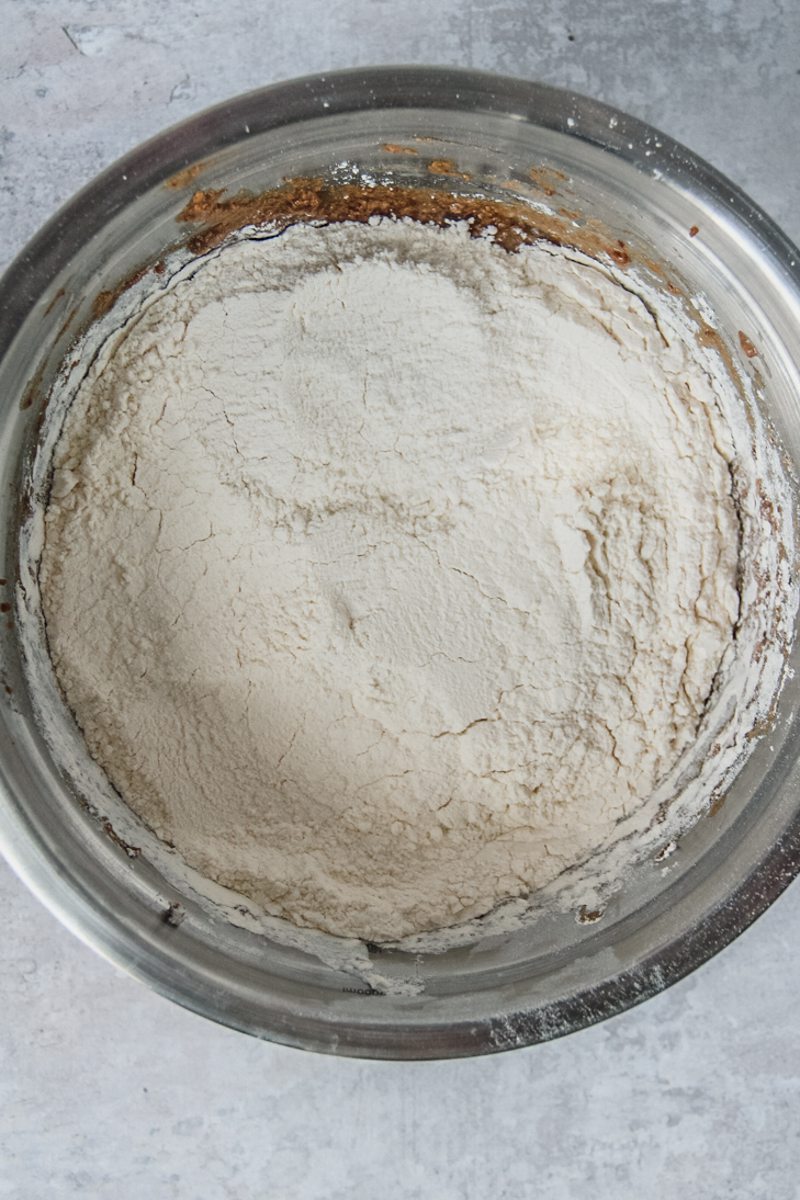 Flour sits on top of a sticky toffee pudding wet mixture in a stainless steel bowl on a gray surface.