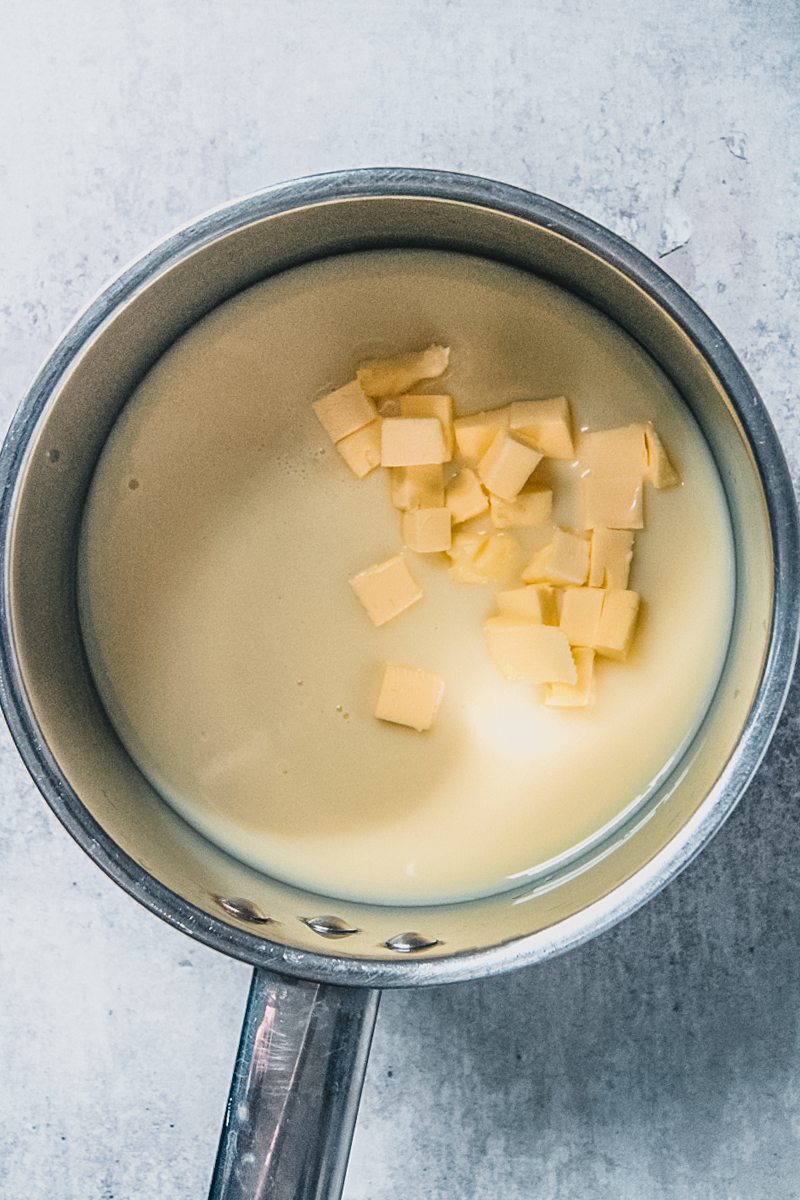 A sweetened condensed and butter mixture sits in a stainless steel saucepan on a gray surface.