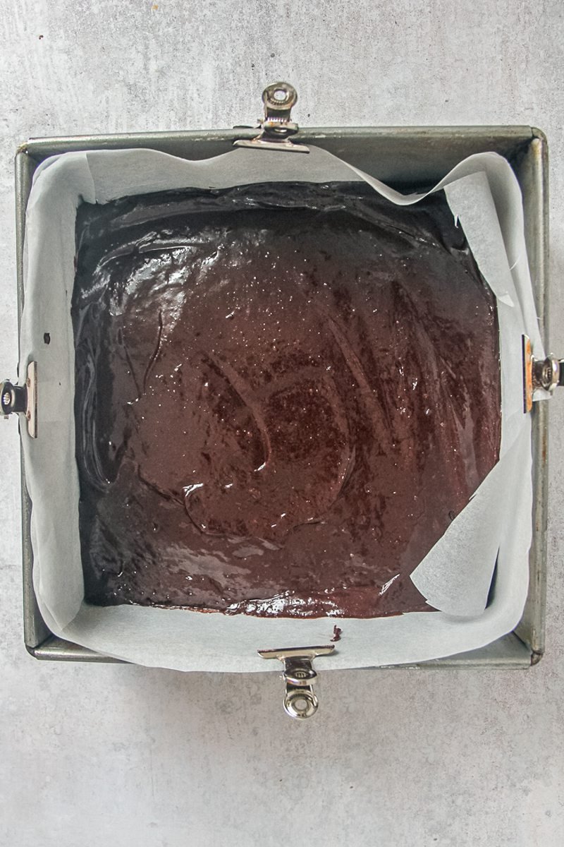 Unbaked brownie sits in a lined square metal baking tin on a gray surface.