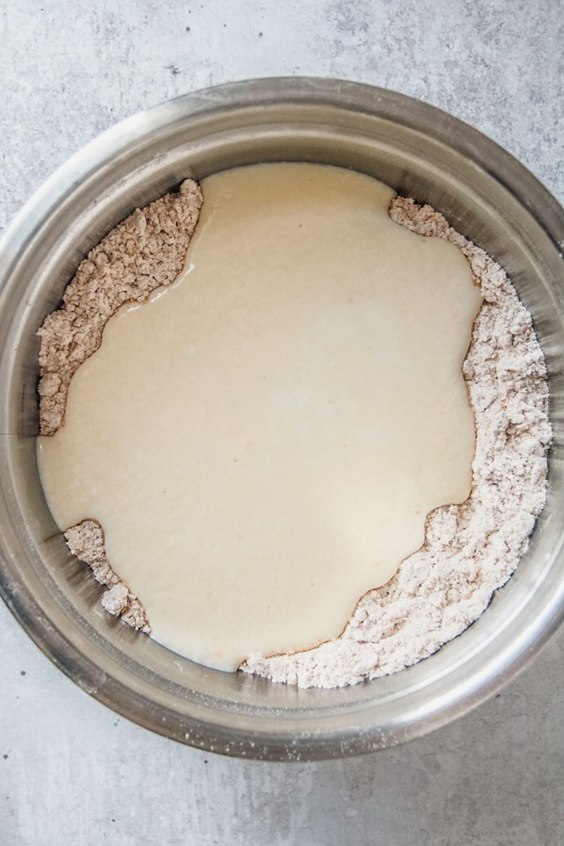 A buttermillk mixture sits in the center of a dry mixture in a stainless steel bowl on a gray surface.