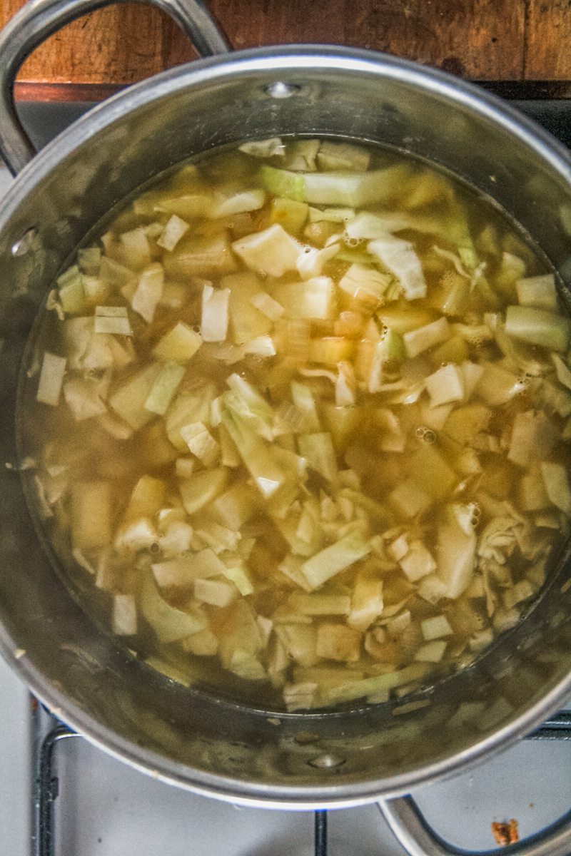 Roughly chopped cabbage and potatoes sit in a broth in a large stainless steel pot on a cooker.