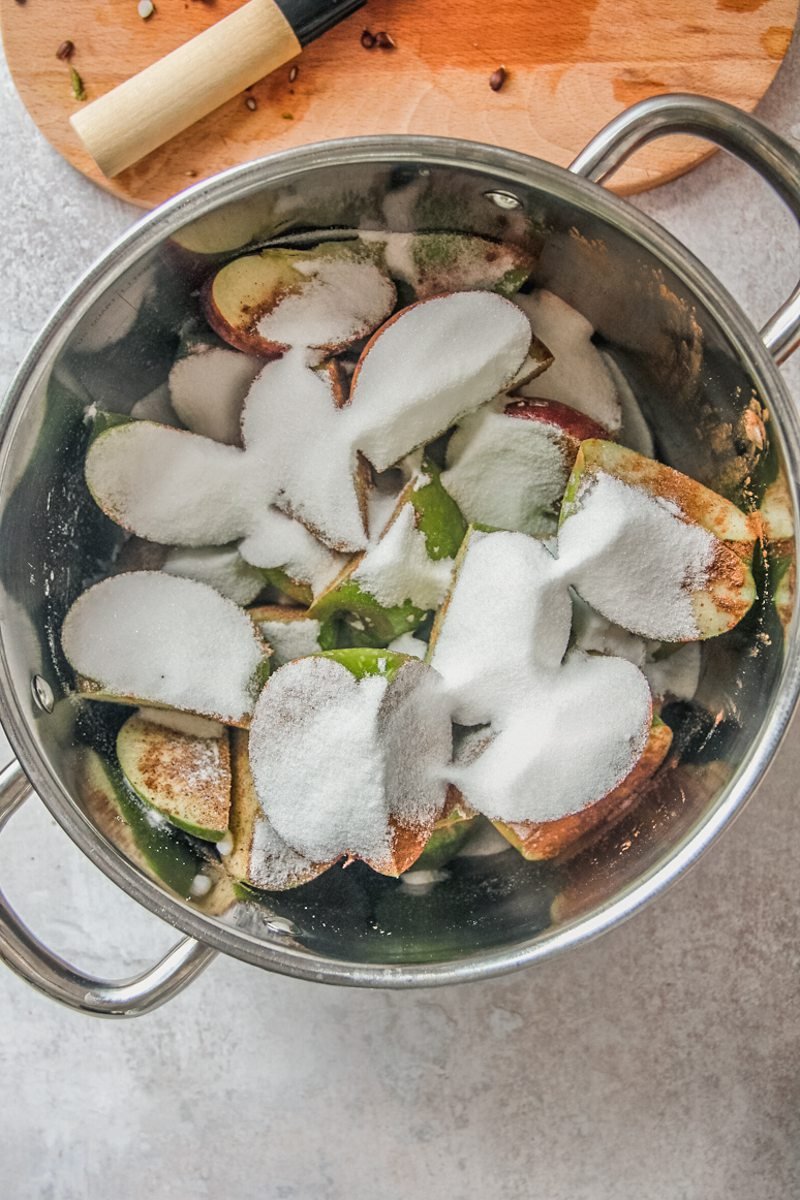 Quartered whole apples sit in a stainless steel pot covered in sugar and spices on a gray surface.