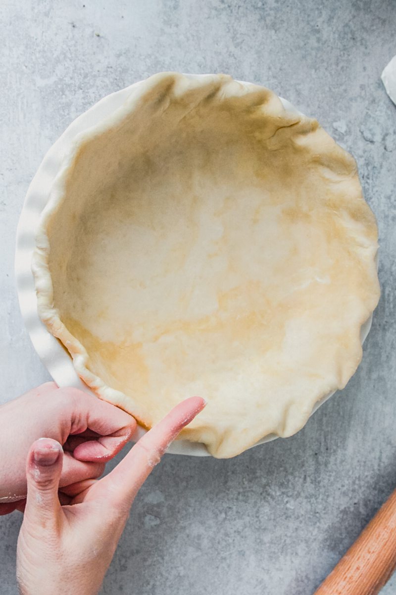 A rope shape is pressed into the edges of a pie dough pressed into a pie dish on a gray surface.