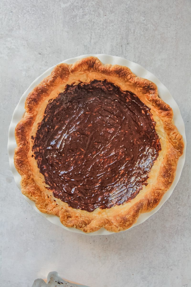 Melted chocolate sits spread over the base of baked pie pastry in a pie dish on a gray surface.