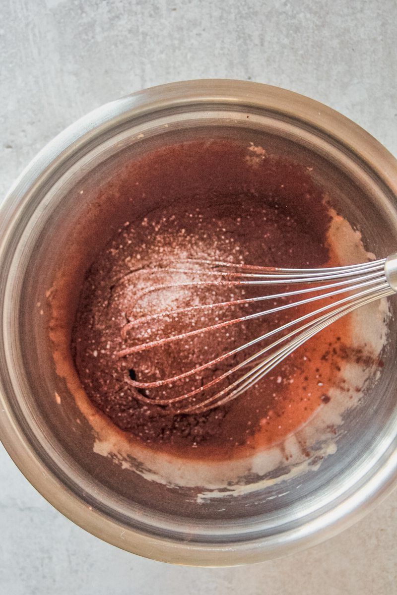 Cocoa powder sits on top of whipped egg yolks with a whisk in a stainless steel bowl on a gray surface.