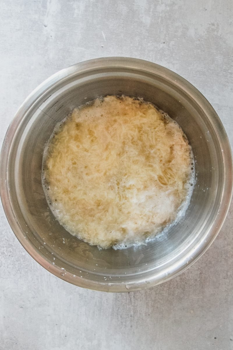 Finely grated potato sits in a stainless steel bowl on a gray surface.