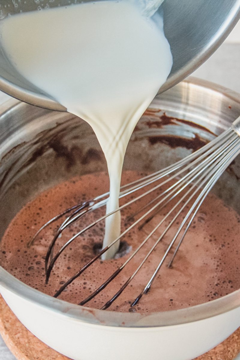 Warm milk is poured into a chocolate mixture in a stainless steel bowl.