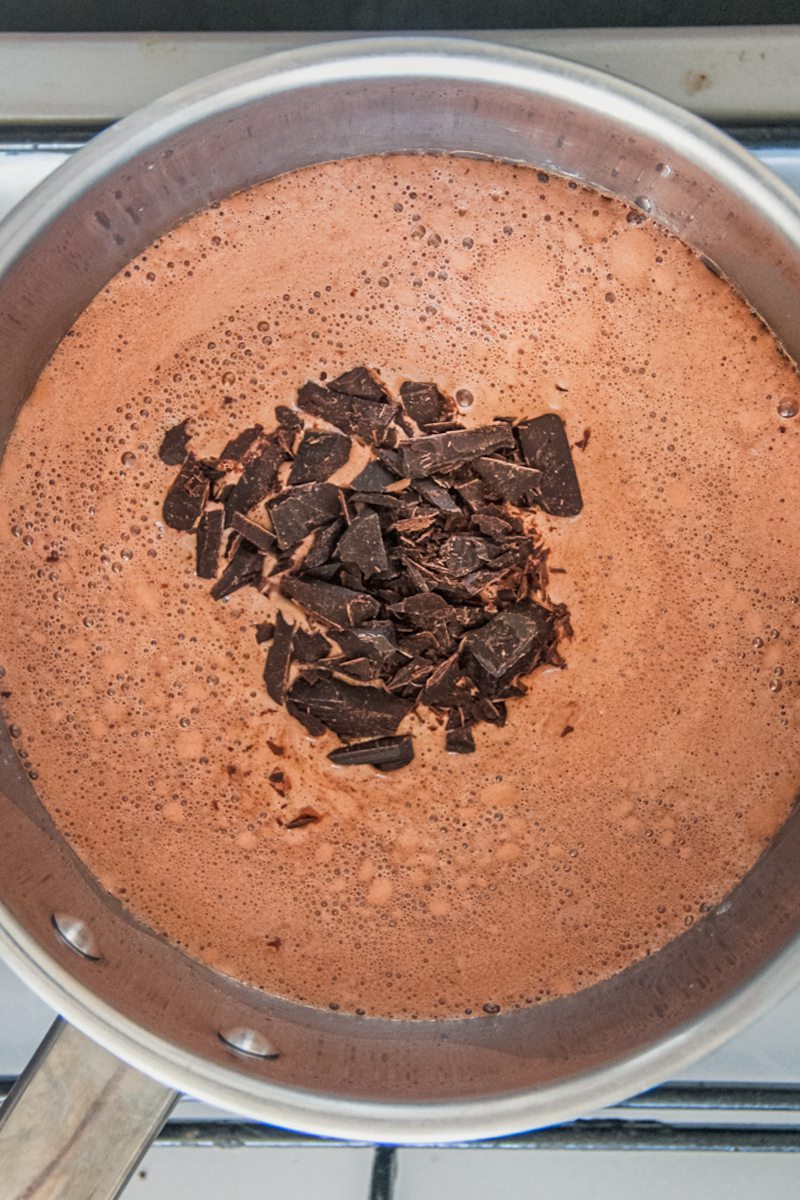 Chocolate chunks sit on top of a chocolate pudding mixture in a stainless steel saucepan on a cooker.