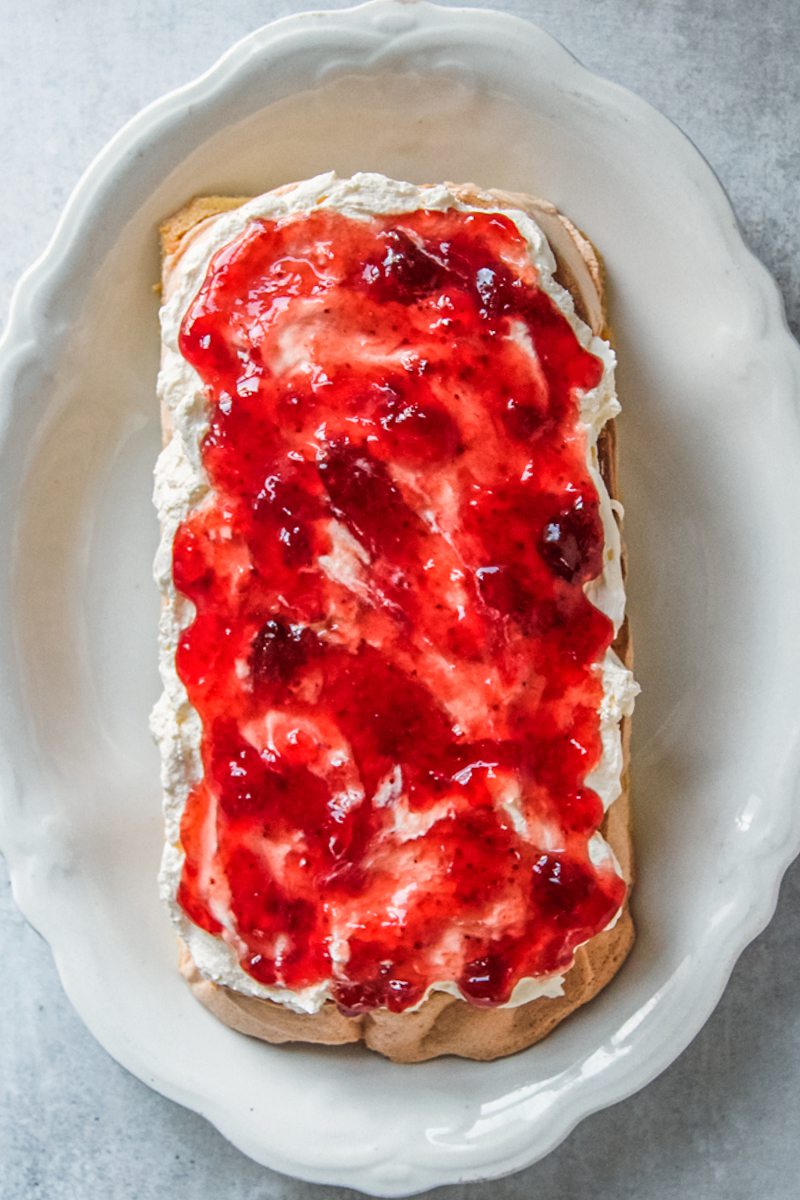 Strawberry jam sits on top of whipped cream on a meringue cake on a white oval ceramic plate on a gray surface.