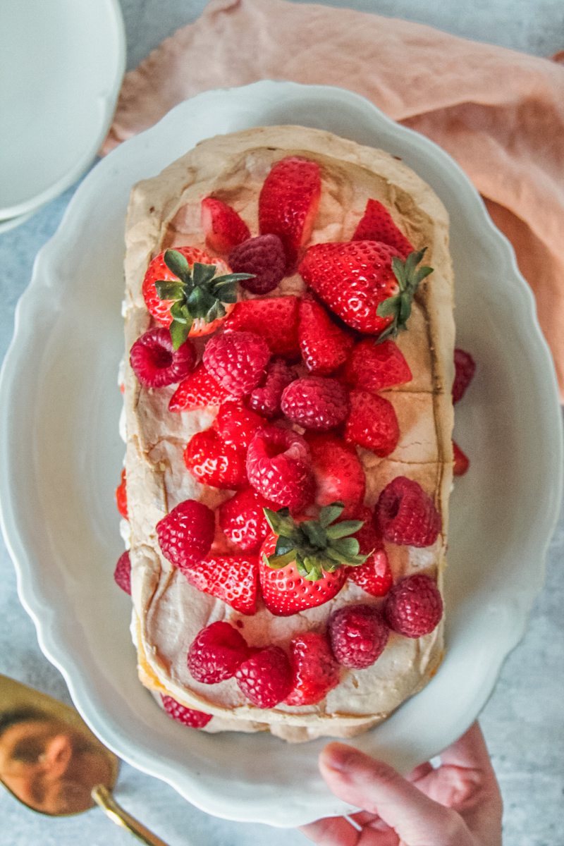 Berries sit atop a meringue cake on a white oval ceramic plate above a gray surface.