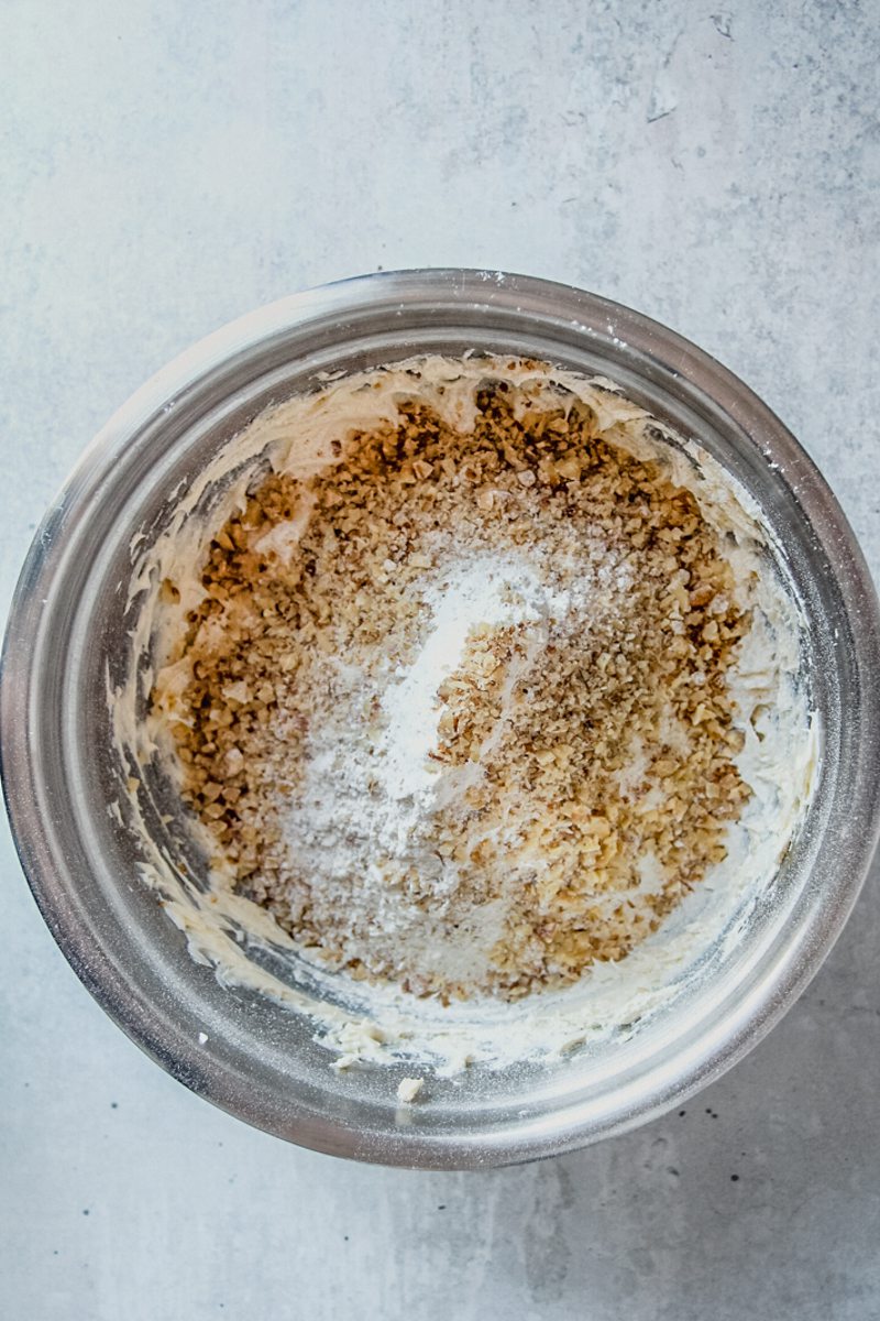 Finely chopped walnuts and flour sit on top of whipped butter and sugar in a stainless steel bowl on a gray surface.