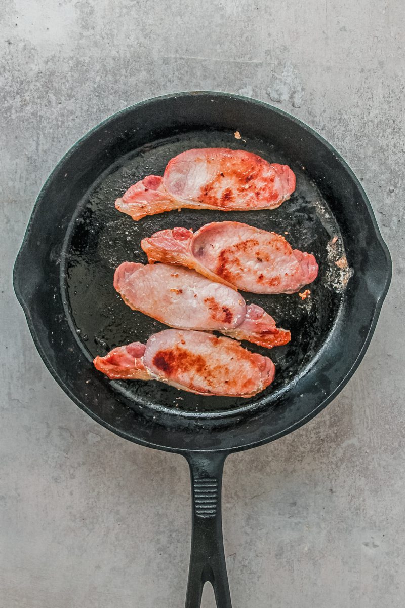 Bacon rashers sit in a cast iron skillet on a gray surface.