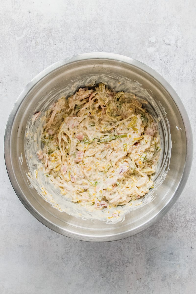 Cooked leeks and bacon sit in a stainless steel bowl with creme fraiche stirred through on a gray surface.