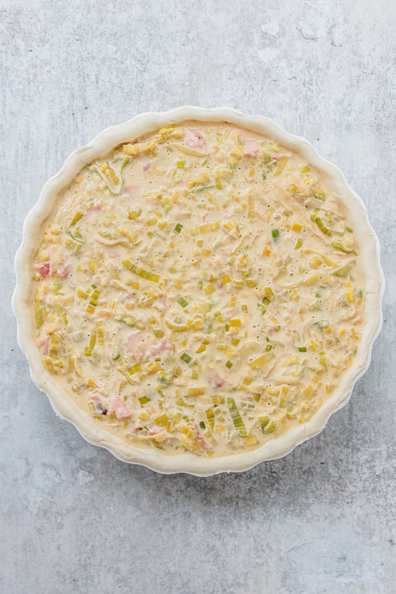 Unbaked Flamiche sits in a tart dish on a gray surface without grated cheese.