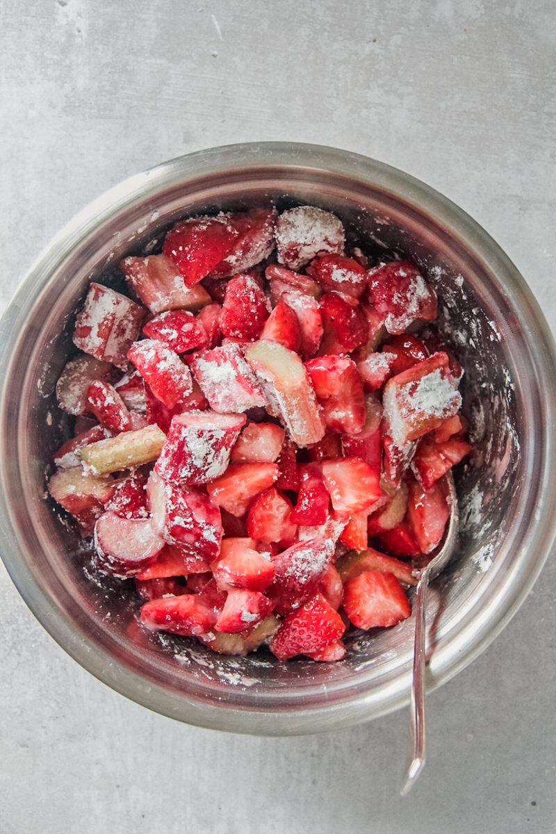 Roughly sliced strawberries and rhubarb sit stirred with cornflour and sugar in a stainless steel bowl on a gray surface.