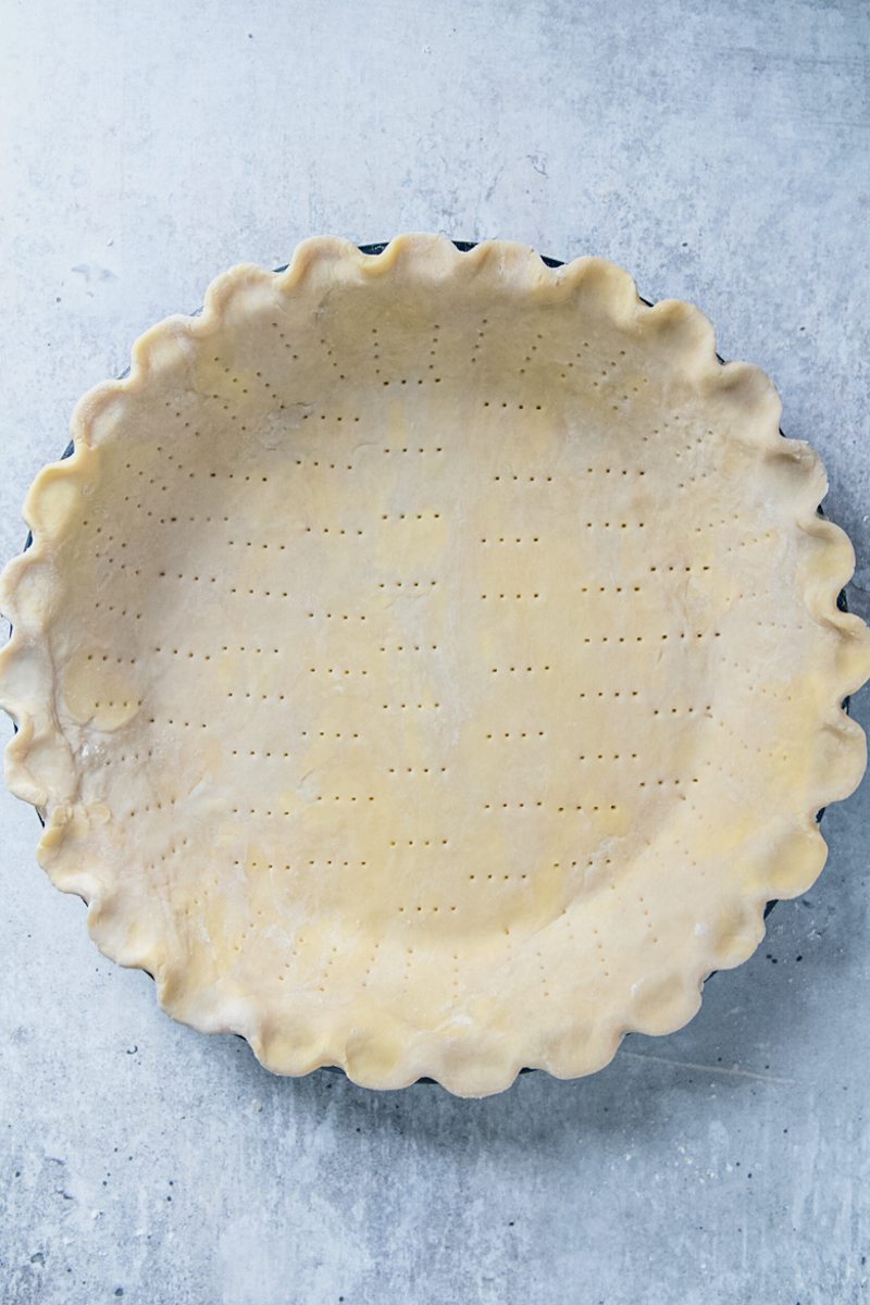 Unbaked pastry is pressed into a pie dish, pierced with a fork and shaped on a gray surface.