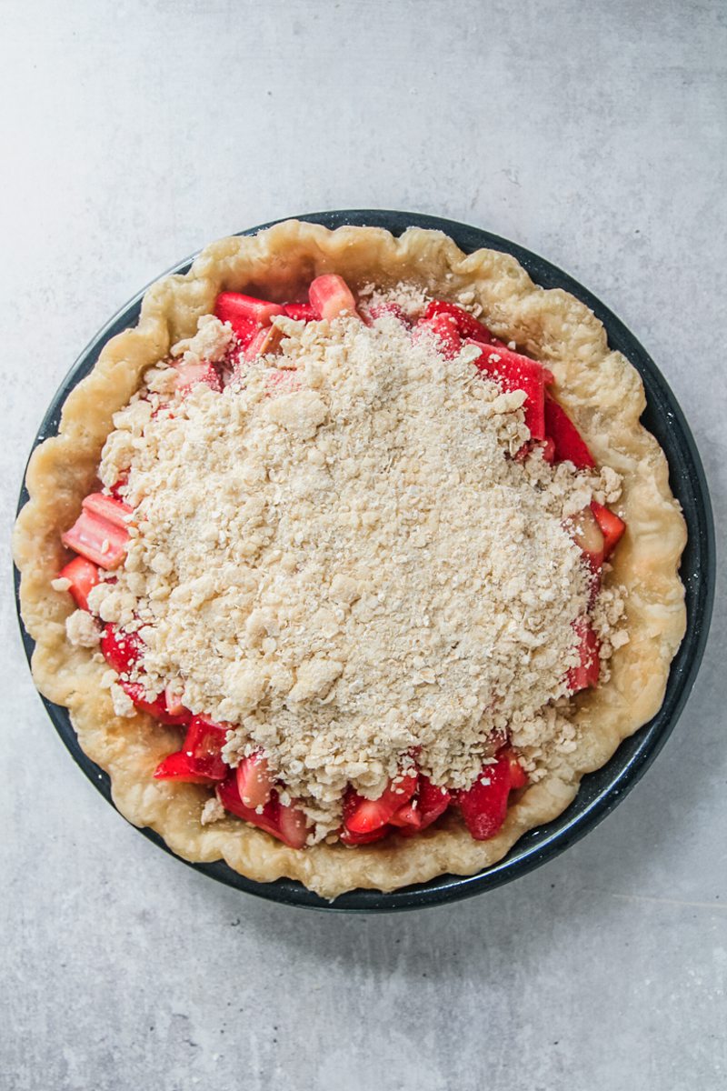 A strawberry rhubarb pie sits parbaked in a black metal pie dish with a streusel topping on a gray surface.