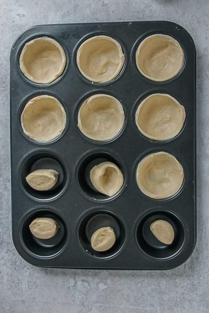 Laminated dough balls sit in a muffin tin, some pressed into the tin and some yet to be pressed; on a gray surface.