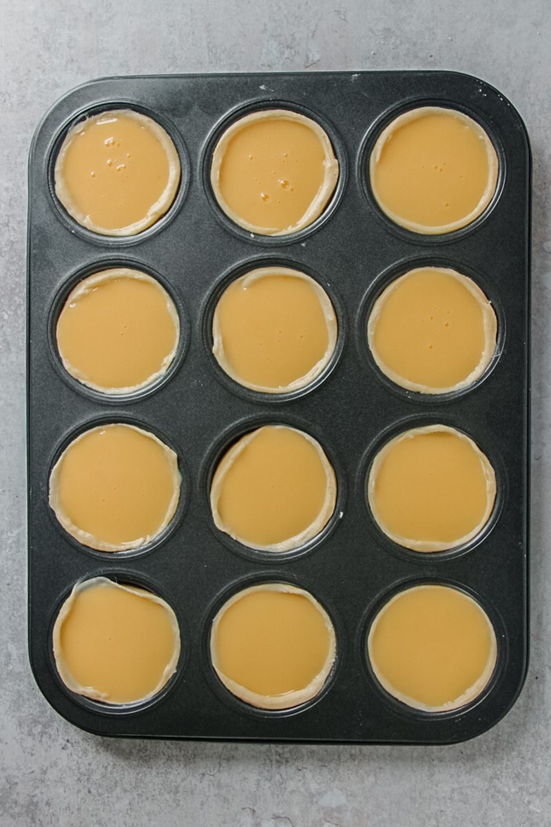 Unbaked Portuguese tarts sit in a muffin tin on a gray surface.