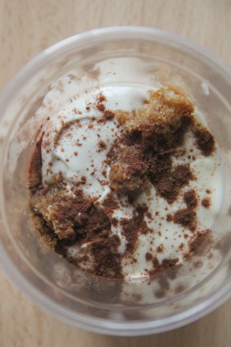 Crumbled coffee soaked sponge fingers sit on top of a custard layer with a sprinkling of cocoa powder in a round plastic cup on a wooden surface.