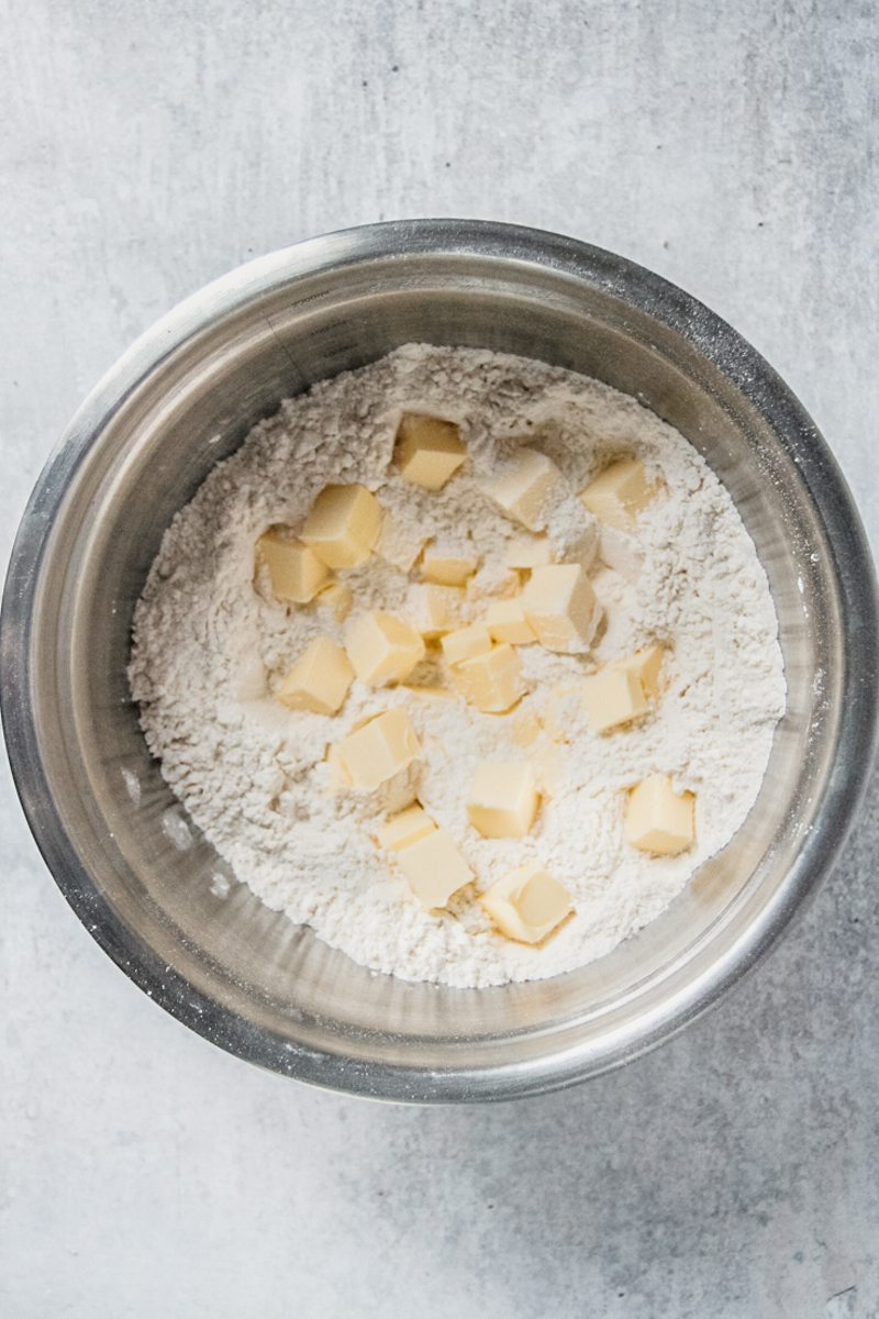 Cubes of butter sit in flour in a stainless steel bowl on a gray surface.