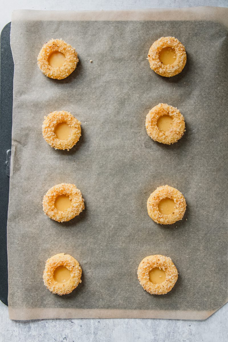 Thumbprint cookies sit on a lined baking tray before the apricot jam filling is placed in the center on a gray surface.