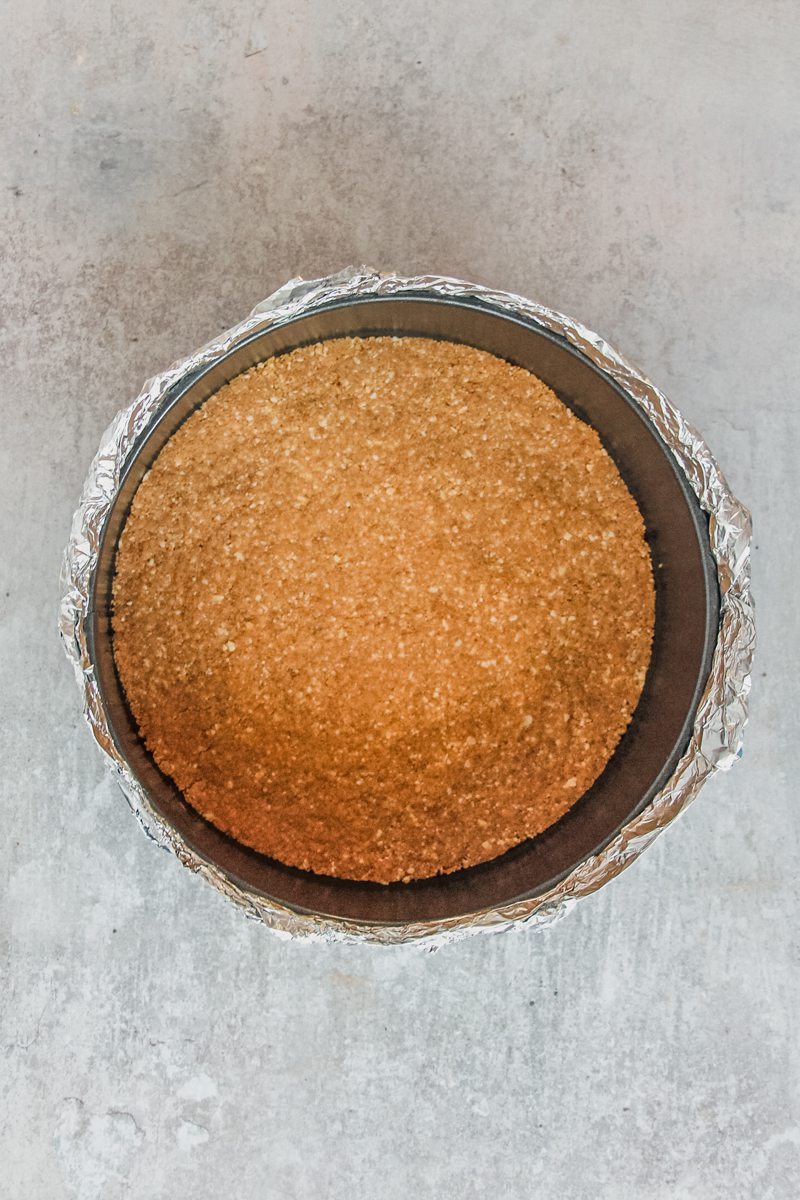 A baked cheesecake base sits in a round metal cake tin with a aluminium foil wrapping.
