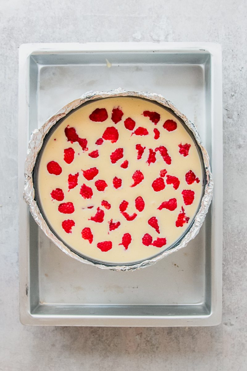 Raspberries sit swirled in a cheesecake mixture in a cake tin wrapped in aluminum foil in a larger tray for a hot water bath to bake in on a gray surface.
