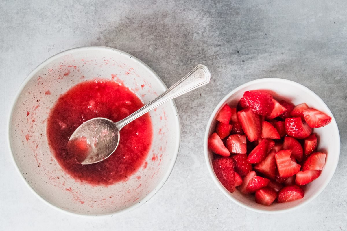 One bowl with mashed strawberries and a second with roughly chopped strawberries sit on a light grey background.