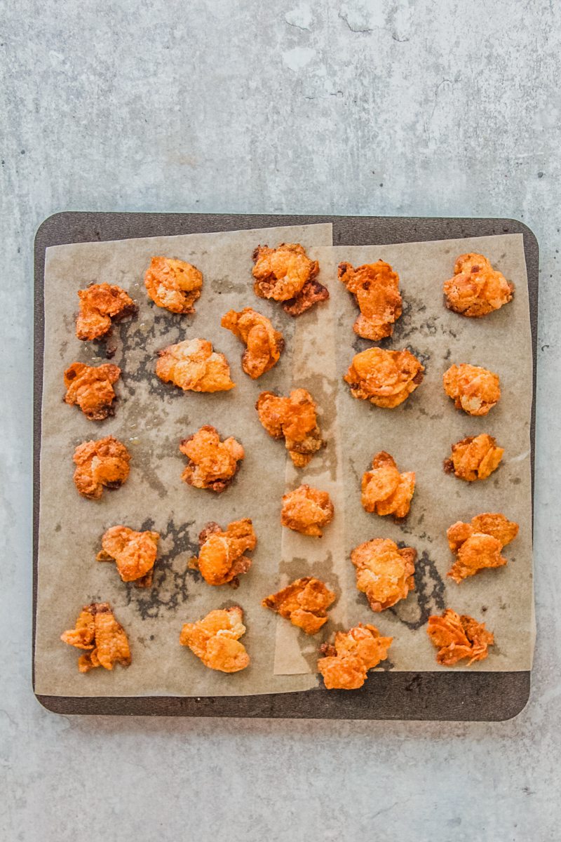 Golden baked cornflake crunch pieces sit on a baking tray on a gray surface.